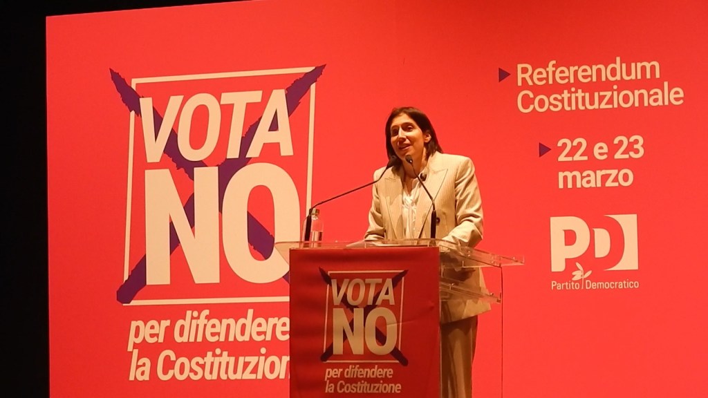 A woman speaking at a podium with a red background displaying 'VOTA NO per difendere la Costituzione' and details about a constitutional referendum in Italy.