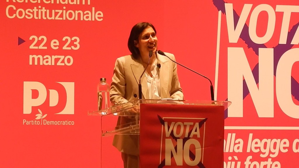 A woman speaks at a podium during an event promoting a constitutional referendum, with a red background displaying 'VOTA NO' and details about the referendum dates.