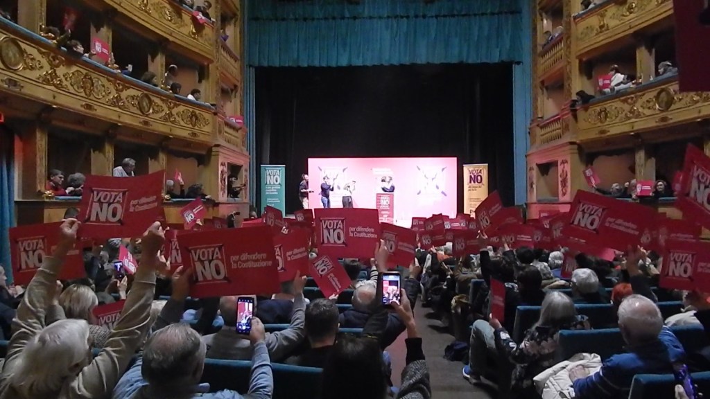 A crowded theater with an audience holding up red signs that say 'VOTA NO' during a public event.
