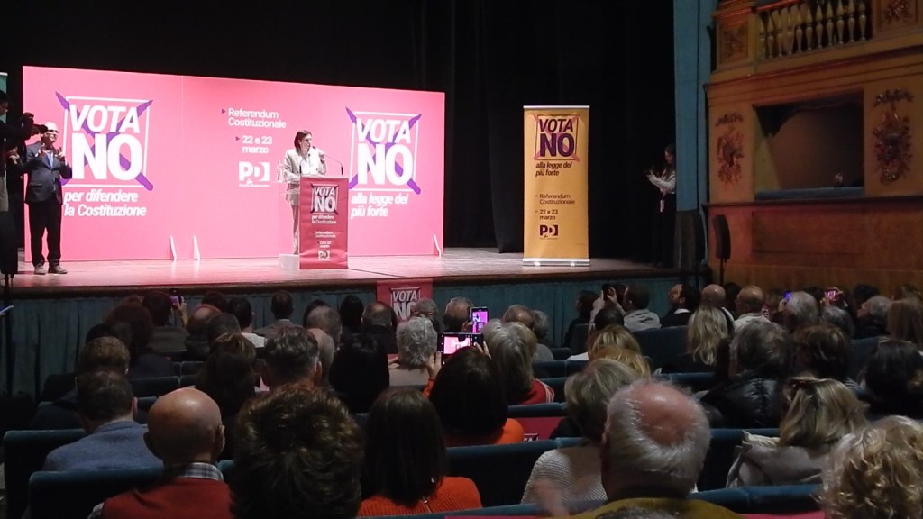 A speaker addresses an audience at a political rally with large 'VOTA NO' banners in the background, advocating against a constitutional referendum.