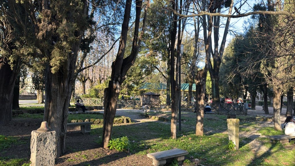 A serene park scene featuring tall trees and stone benches, with people relaxing in the background and a fence visible.