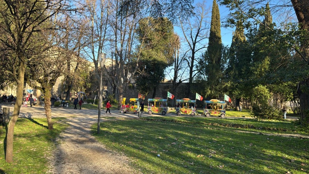 A scenic park pathway with trees, people walking, and a colorful tourist train decorated with flags.