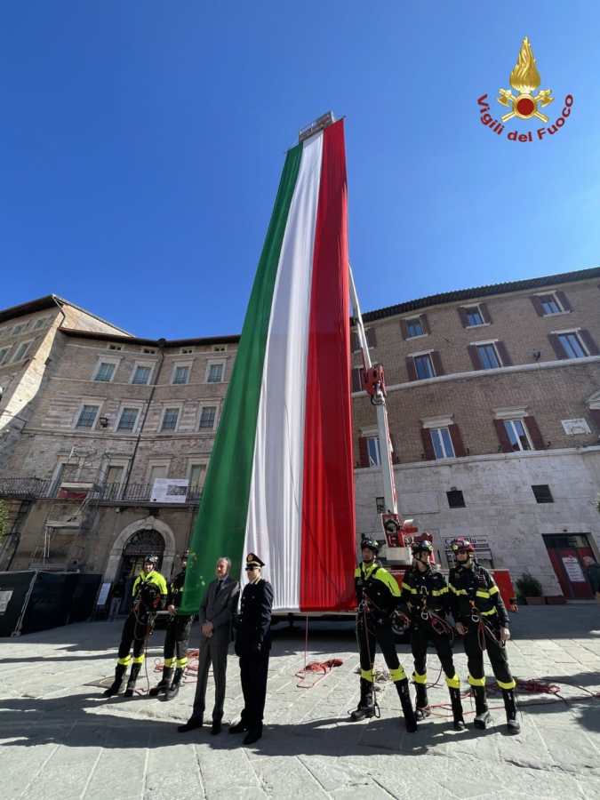 A large Italian flag displayed prominently with several individuals, including firefighters, standing in front. The scene is set in a square with historic buildings.