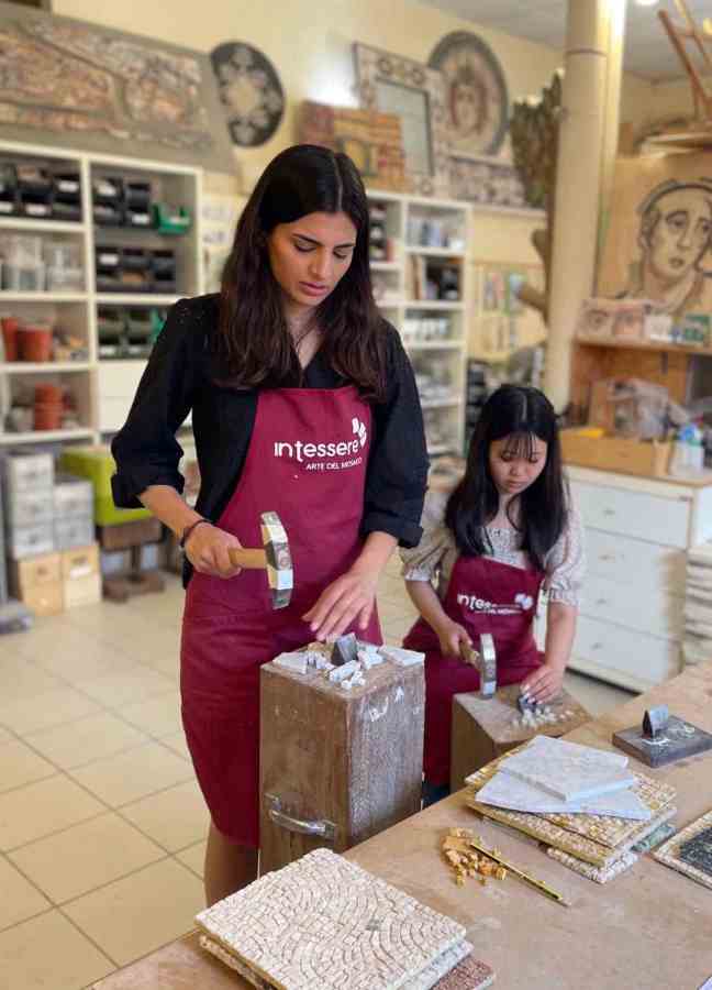 Two women working on mosaic art in a studio; one woman is hammering stone pieces while the other is seated, arranging materials.