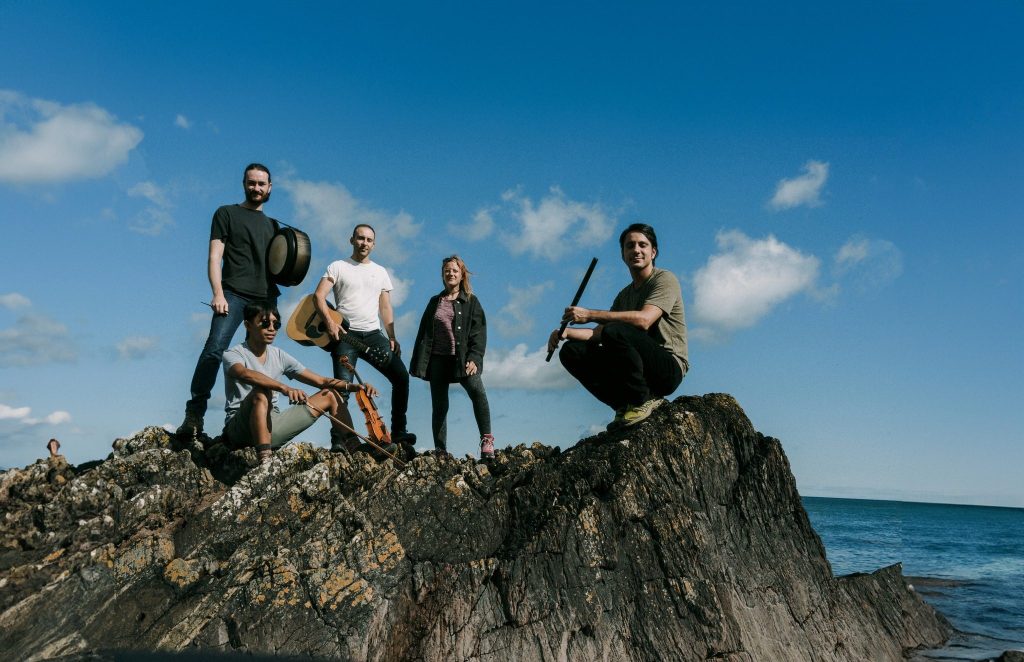 A group of five people posing on a rocky shore with the ocean in the background, under a blue sky with scattered clouds. Two individuals are sitting, while three are standing, showcasing casual clothing and musical instruments.