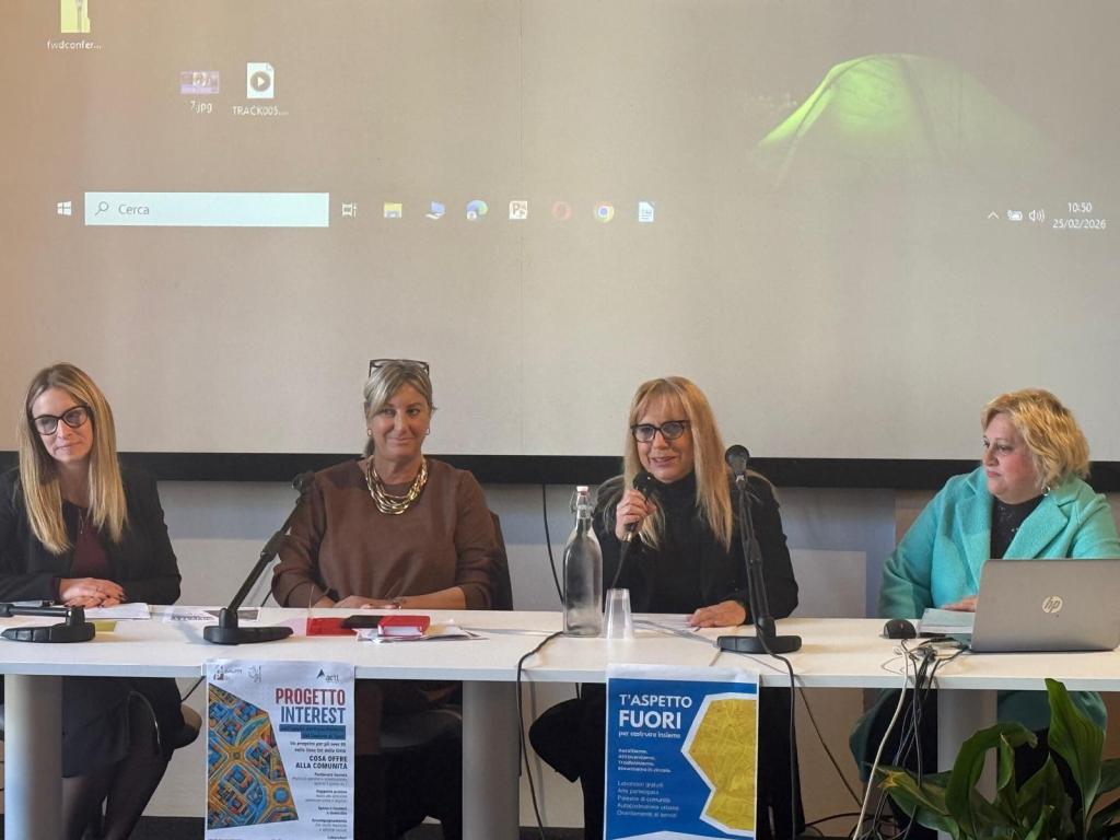A panel discussion featuring four women seated at a table with microphones, discussing a project. A large screen is visible in the background displaying icons. Pamphlets are placed on the table, and a glass bottle and glasses are present.