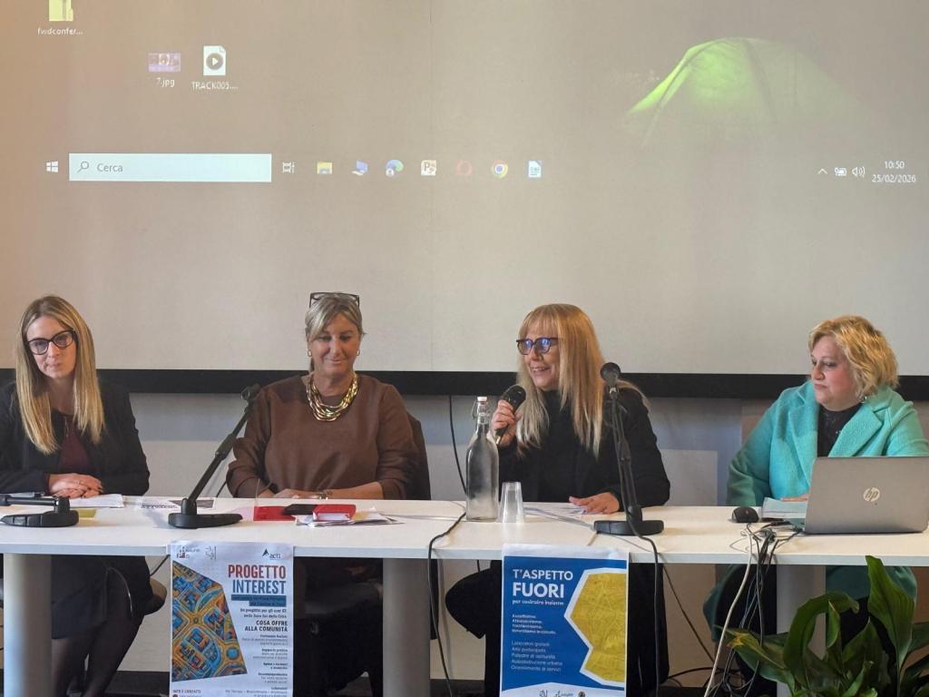 A conference panel featuring four women seated at a table with microphones, discussing topics related to a community project, with a projector screen displaying a desktop background.