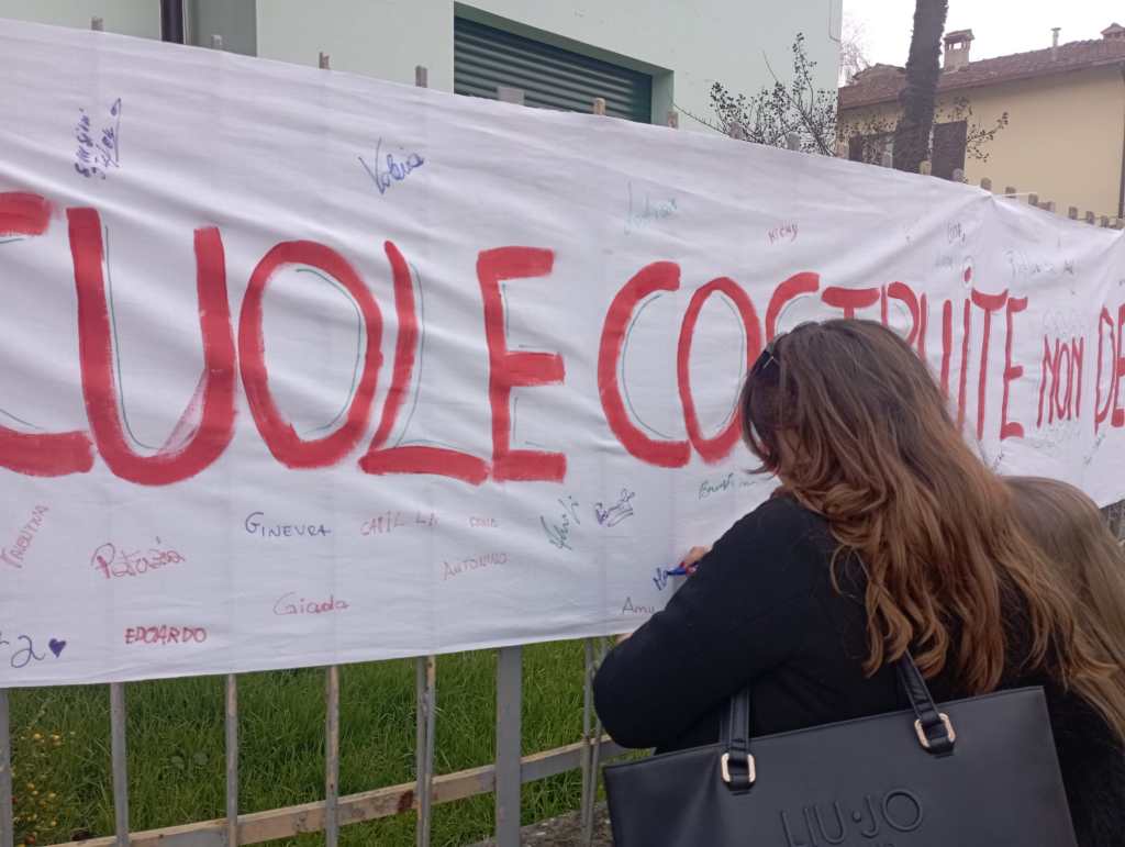 A woman stands by a large banner that reads 'CUOLE COSTRUITE NON DEBILITATE,' decorated with various signatures and drawings. The setting appears to be a community gathering or protest related to education.