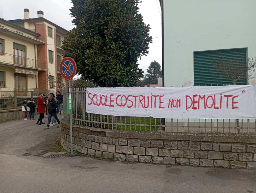 A banner reading 'SCUOLE COSTRUITI NON DEMOLITE' hung on a fence, with a group of people walking nearby. The setting features residential buildings and a no parking sign.