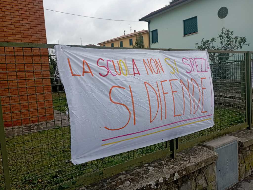 A large white banner hanging on a fence that says 'LA SCUOLA NON SI SPEZZA SI DIFENDE' in colorful letters.