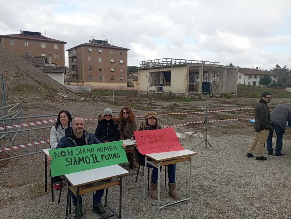 Protesters seated at desks holding signs with messages in Italian, expressing their concern about being viewed as mere numbers while advocating for a future. Background features construction and empty land.