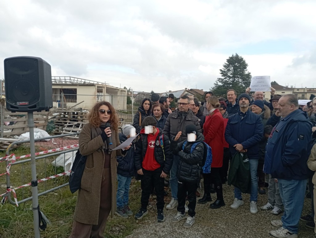 A woman speaks at a rally, holding a microphone, with a crowd of people behind her. Some children stand beside her. In the background, there is construction material and a cloudy sky.