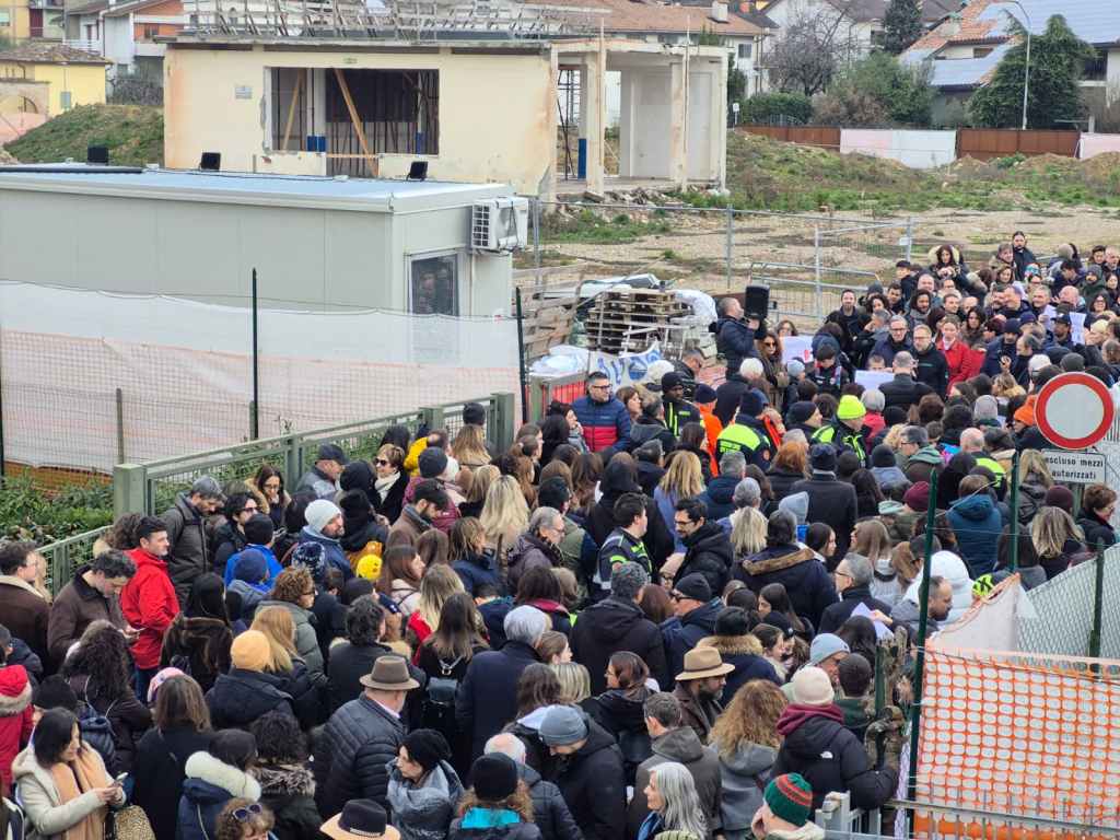 A large crowd of people gathered outdoors near a construction site, with some participants wearing safety vests. A temporary structure is visible in the background alongside unfinished buildings.