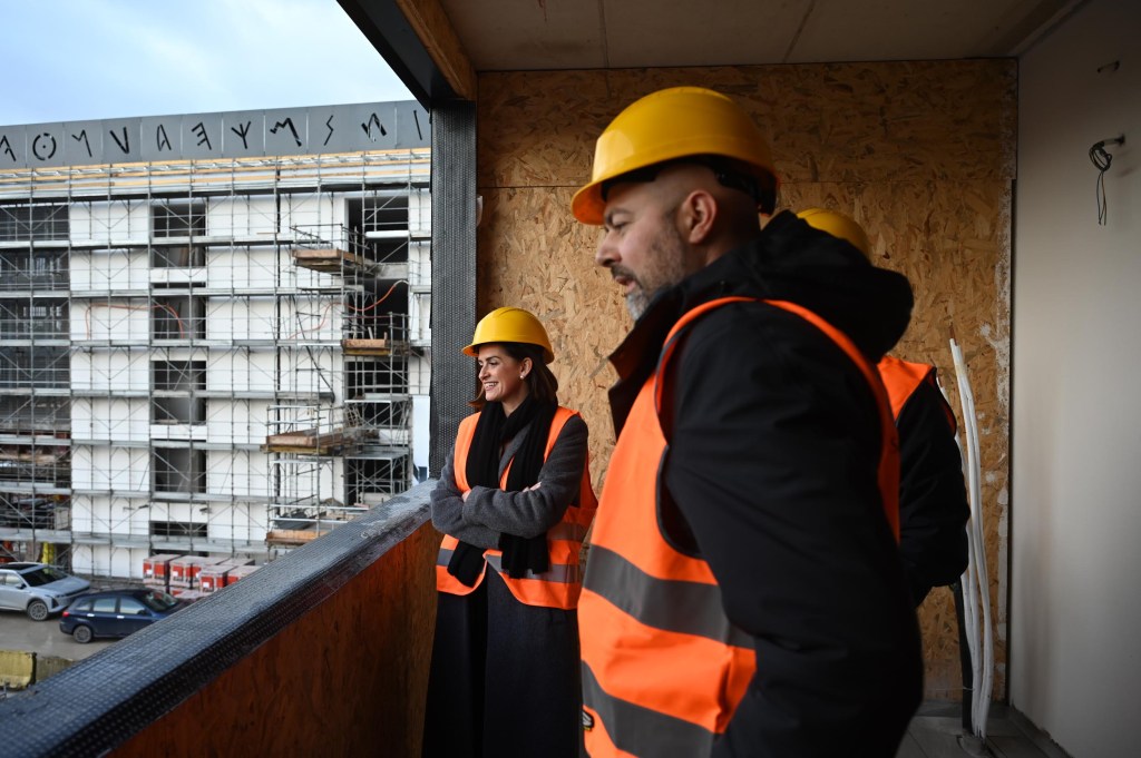 A group of construction professionals, including a woman and men, wearing safety helmets and vests, stands on a balcony overlooking a building site with scaffolding in the background.