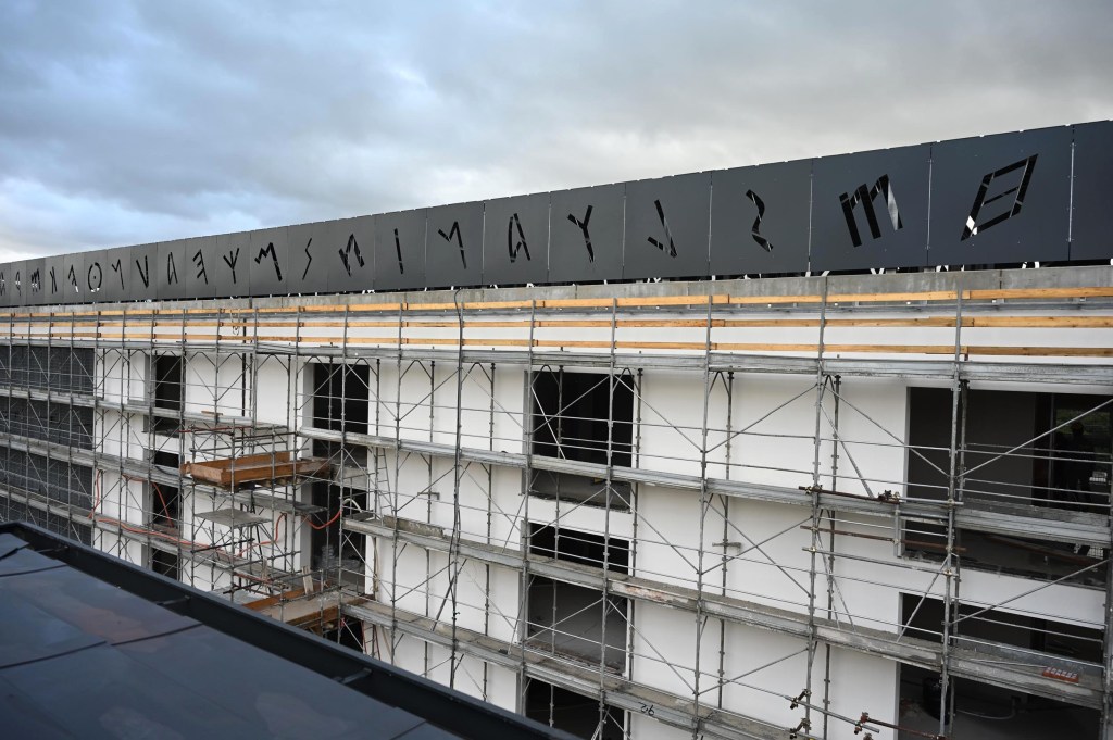A construction site featuring a partially completed building with scaffolding and a black panel displaying cut-out letters on the roof.