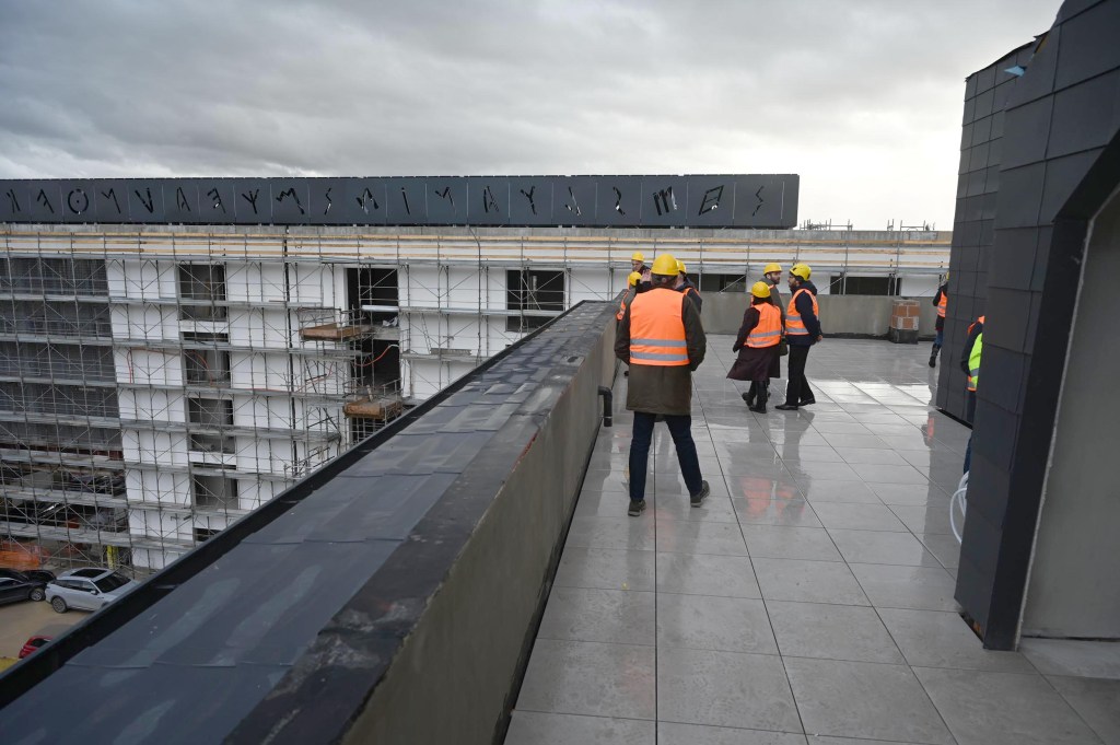 Construction workers wearing hard hats and safety vests on a rooftop during a site visit, with a partially constructed building and scaffolding in the background.