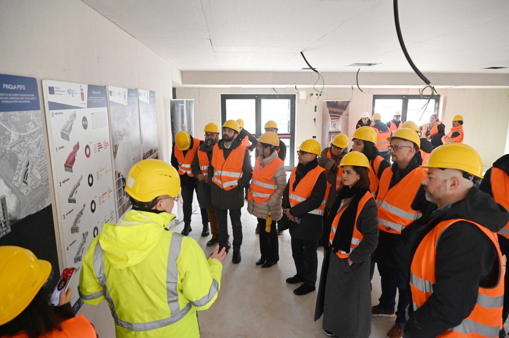 A group of visitors wearing yellow hard hats and orange safety vests listens to a presentation inside a construction site, with informational posters on the walls.