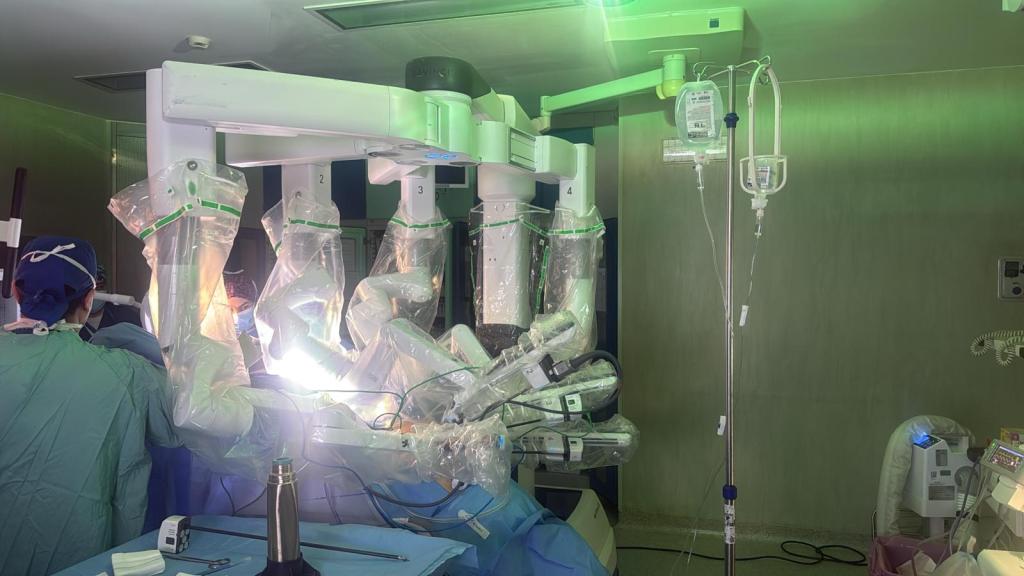 A modern robotic surgical system with multiple articulated arms covered in sterile drapes, positioned over a surgical table in an operating room. Medical staff in scrubs and surgical caps are visible in the background, and there are IV bags and medical equipment present.