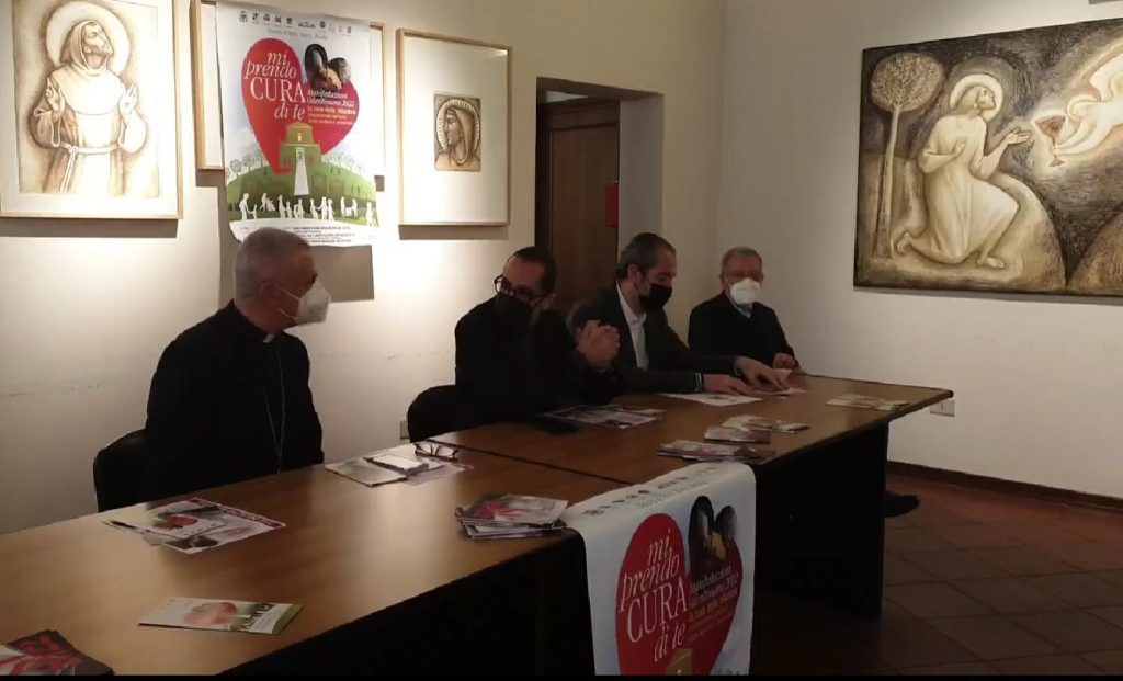 Four individuals seated at a table in a conference room, engaging in a discussion while wearing masks. Behind them, there are framed artworks on the walls and a poster featuring a heart design promoting an initiative.