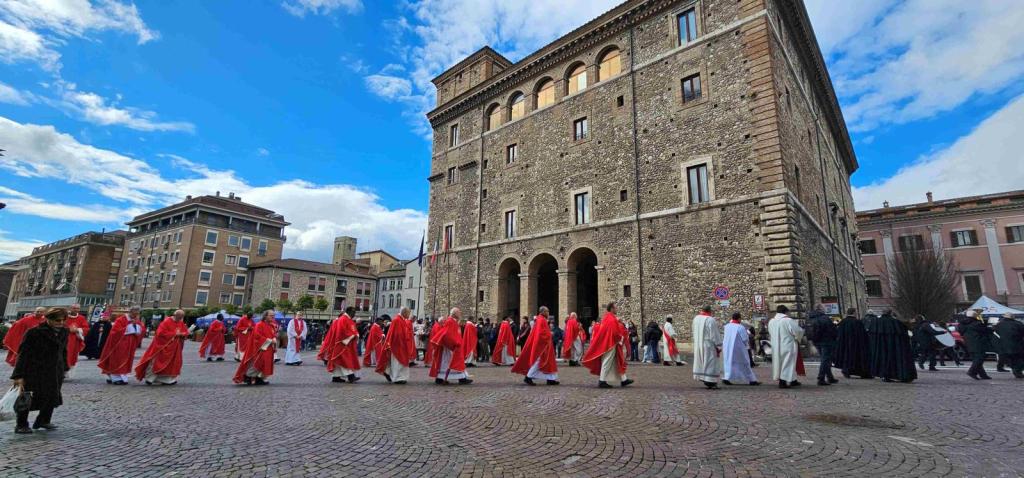 A procession of clergy in red robes walking in front of a historic stone building under a partly cloudy sky.