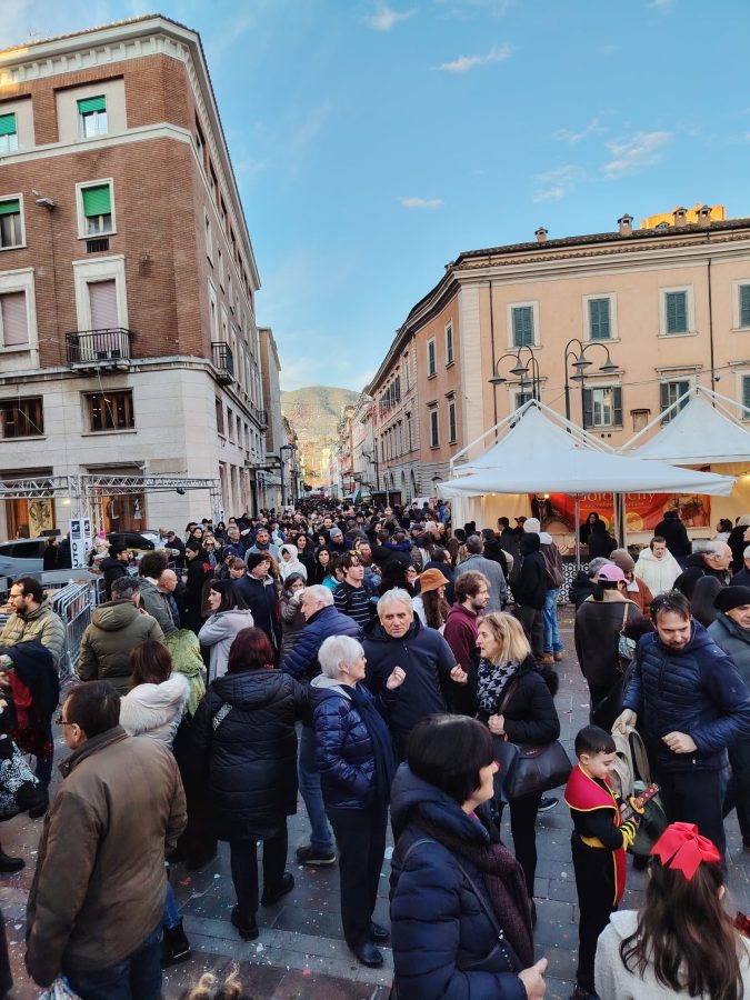 A crowded street scene with many people gathered, some interacting and others looking around, alongside buildings and white tents set up for an event under a clear blue sky.