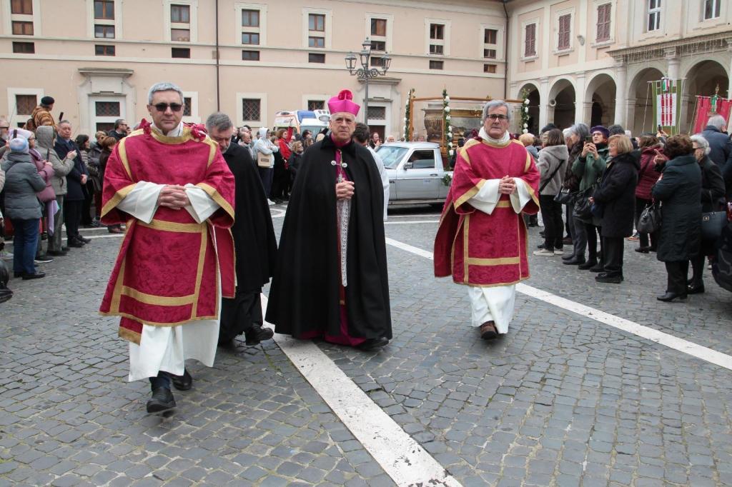 Three clergy members dressed in red robes and a black cloak walk along a cobblestone street during a religious ceremony, surrounded by a crowd of onlookers.