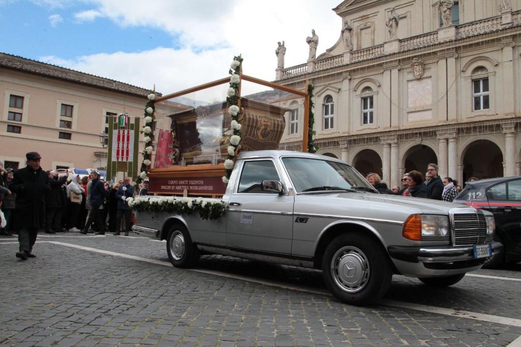 A silver pickup truck is transporting a glass-enclosed display with flowers, showcasing a statue or relic, during a procession in a city street. A crowd of people watches from the sidewalk, with buildings and a blue sky in the background.