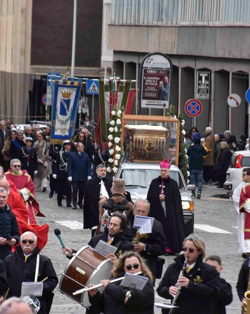 A religious procession featuring a bishop, a decorated vehicle carrying a religious artifact, and participants in traditional attire, marching through a street with onlookers and banners.
