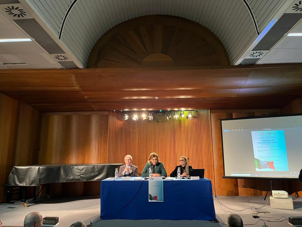 A conference scene featuring three speakers at a blue table, with a presentation on a screen behind them. The room has wooden paneling and spotlighting.