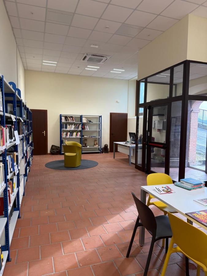 Interior view of a modern library featuring bookshelves filled with books, a circular yellow chair in the center, and a white table with chairs. The flooring is terracotta tiles and there are large windows allowing natural light.