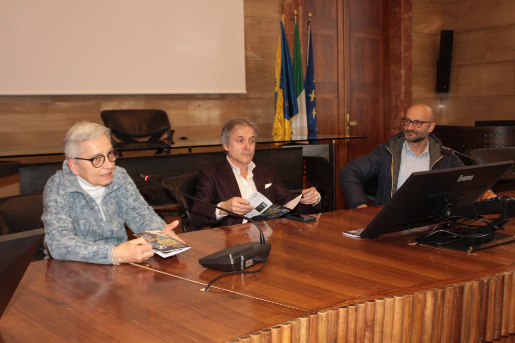 Three people seated at a conference table in a formal setting, with a computer monitor in front of them. They appear to be engaged in a discussion, holding brochures. The background includes flags and a presentation screen.