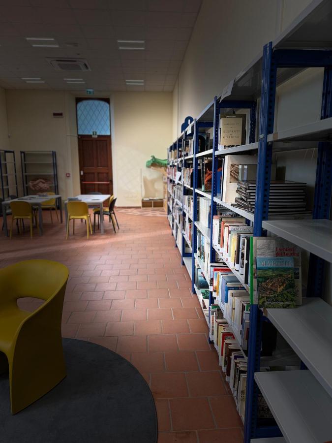 A library interior featuring bookshelves along a corridor, with a round carpet and a yellow chair in the foreground. Tables and chairs are set up for reading or studying, and a wooden door with a window is visible in the back.
