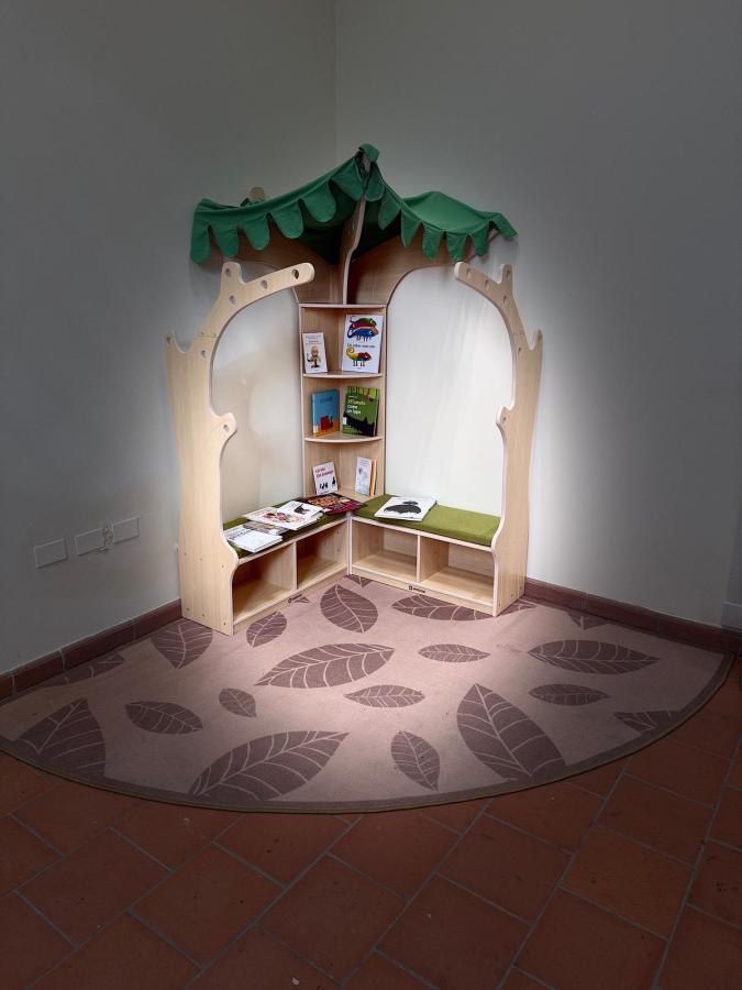 A wooden reading nook designed like a tree, featuring a green canopy and shelves filled with colorful children's books, set against a leaf-patterned floor.