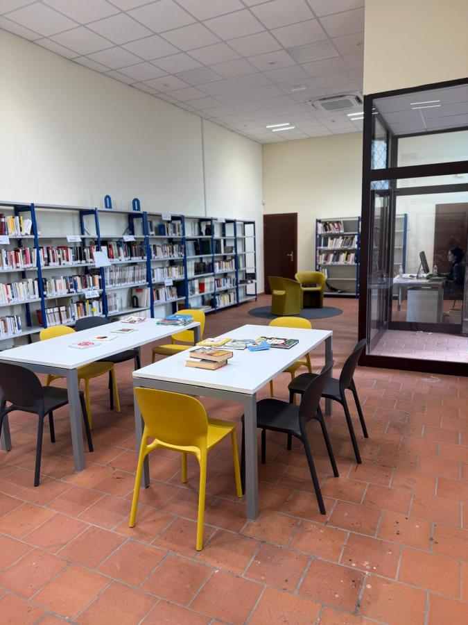 Interior of a library featuring bookshelves filled with books, tables with chairs in yellow and black, and a study area in the background.