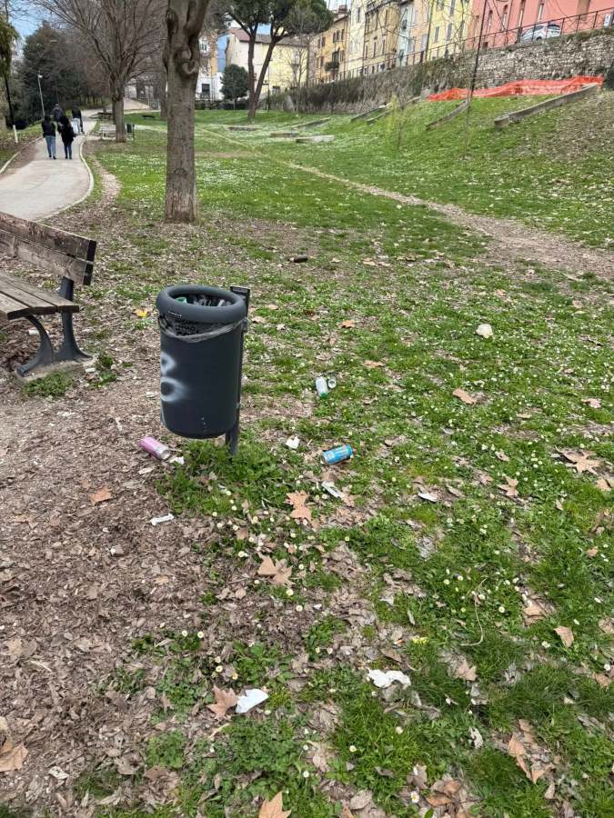 A trash can in a park surrounded by scattered litter and fallen leaves, with a paved path and trees in the background.
