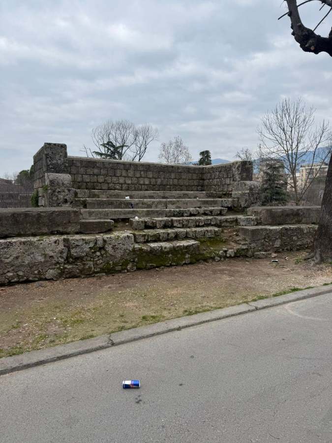 An old stone structure with stepped platforms, surrounded by trees and a cloudy sky, located next to a road. A can is visible on the ground in the foreground.