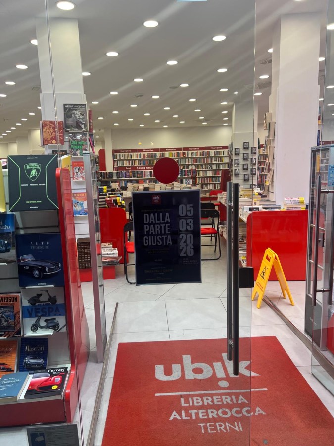 Entrance of Ubik bookstore in Terni, showcasing bookshelves filled with books, a red carpet with the store name, and a caution sign inside.