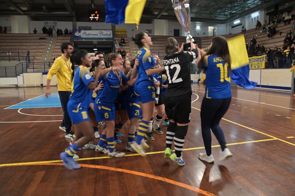 A group of female athletes celebrating a sports victory, holding a trophy and flags, in a gymnasium. The players are wearing blue and yellow uniforms, and the atmosphere is joyful and energetic.