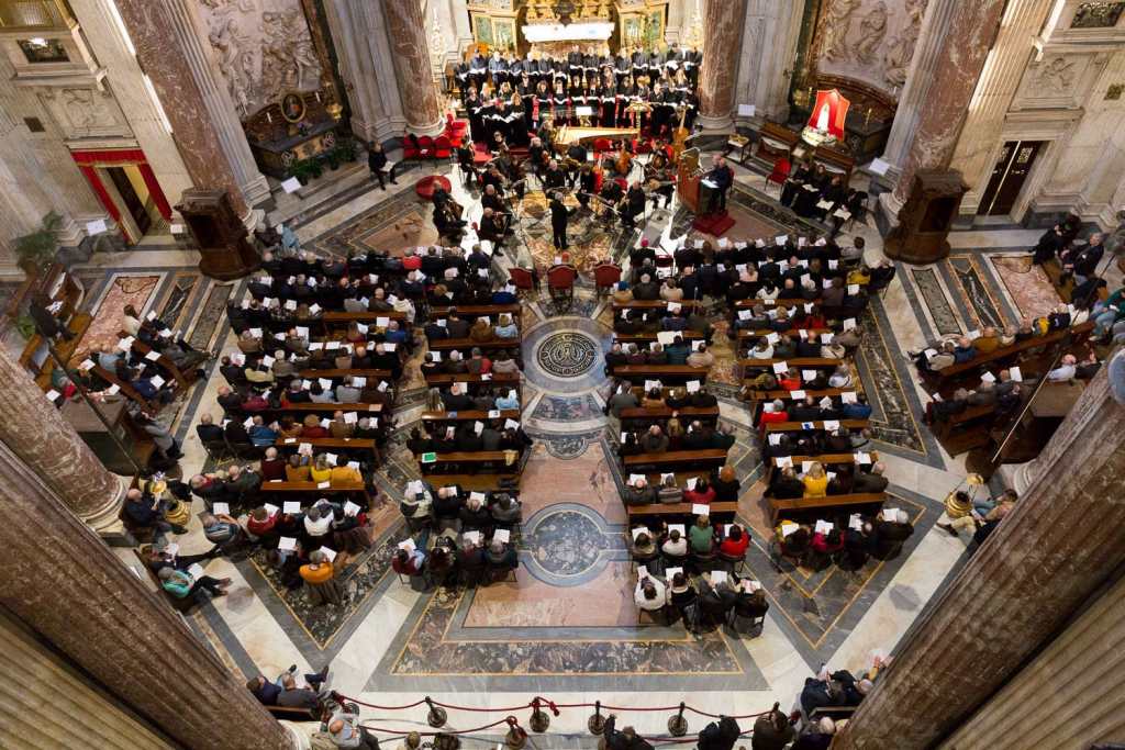 An aerial view of a large gathering in a cathedral, featuring rows of seated attendees facing a choir performing at the front, with ornate architectural details and decorations visible in the background.