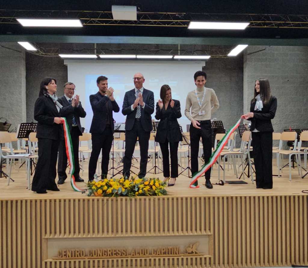 A group of six individuals stands on a stage at a conference center, celebrating a ribbon-cutting ceremony. They are clapping and smiling, with one person holding a ribbon in the colors of the Italian flag. A backdrop of musical instruments and chairs is visible behind them.