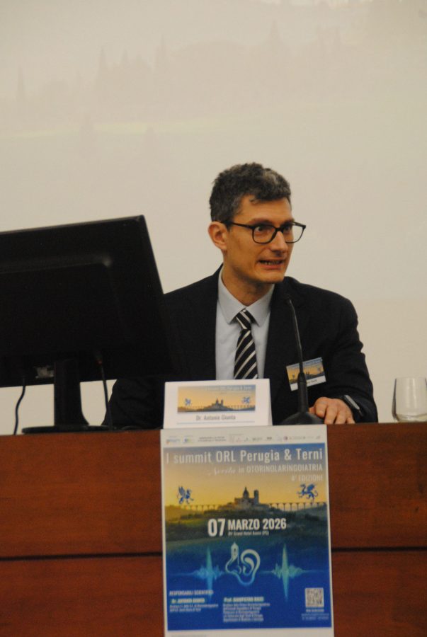 A man in a suit with glasses speaks at a podium during a conference. A monitor is visible beside him, and a colorful event poster for the 'ORL Summit Perugia & Terni' is displayed in front.