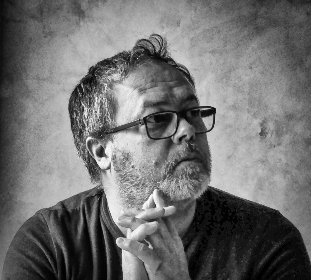 A thoughtful man with glasses and a beard, resting his chin on his hands, against a textured background in black and white.
