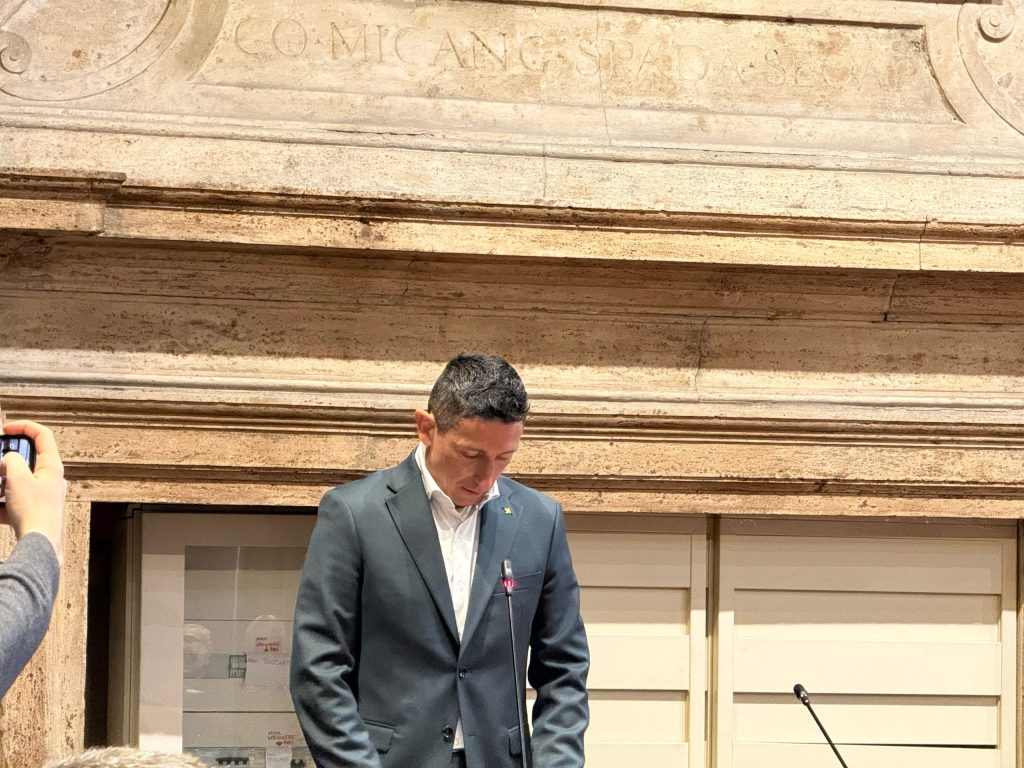 A person in a suit standing at a lectern, looking down, with a stone wall featuring inscriptions in the background.