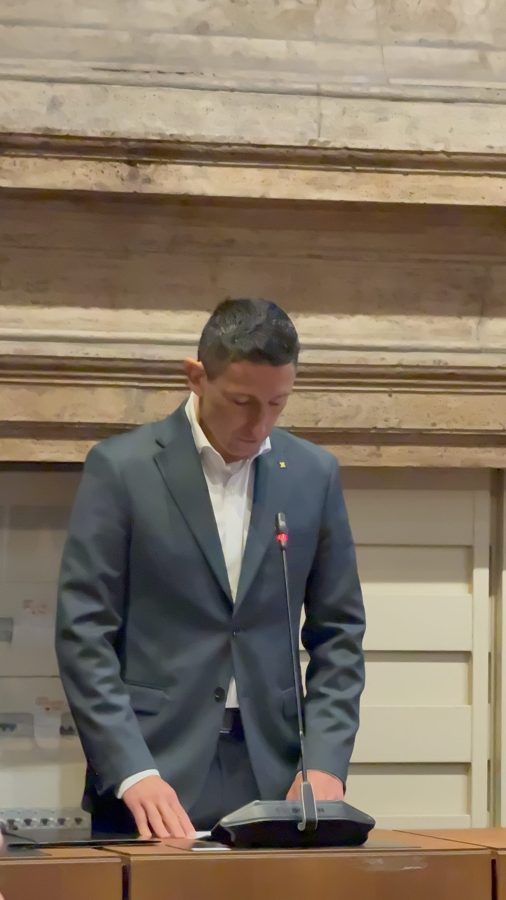 A man in a suit standing at a podium with a microphone, appearing to read or speak, in a formal setting.