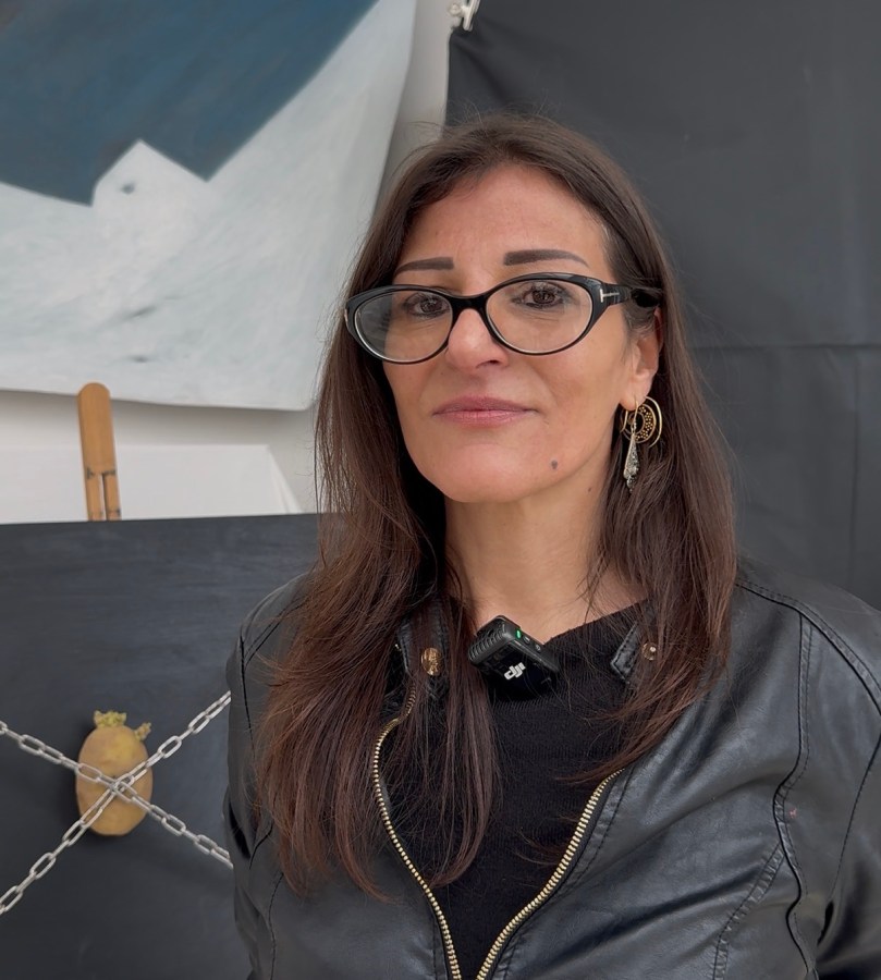 A woman with long brown hair wearing large glasses and a black leather jacket stands in front of a black backdrop with a chain and an object resembling a potato.