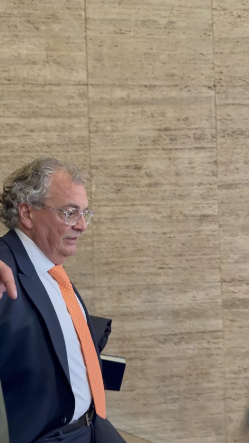 A man with curly gray hair wearing glasses and an orange tie, walking past a textured wall.