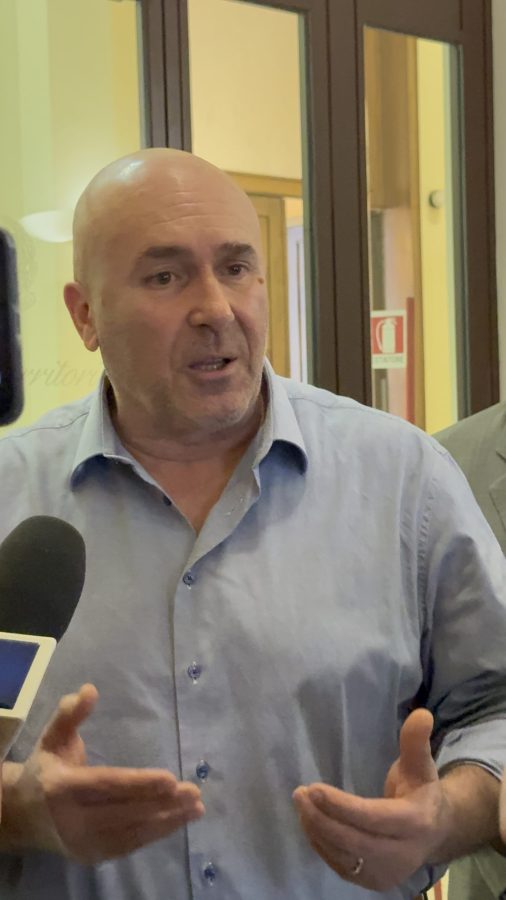 A man with a bald head passionately speaking during a press conference, gesturing with his hands while surrounded by reporters holding microphones.