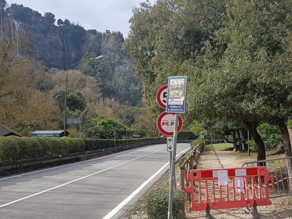 A street scene showing a road with traffic signs, a bus stop, and a construction barrier. In the background, a hillside features a waterfall and trees.