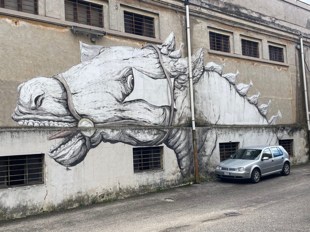 A large, detailed mural of a mythical creature resembling a dragon or a horse painted on a gray wall, with a parked silver car in front.