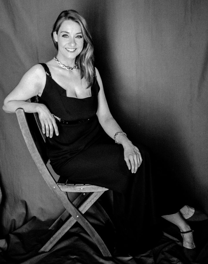 A woman sitting on a wooden chair, wearing a black dress with a low neckline and elegant jewelry, smiling at the camera against a dark backdrop.